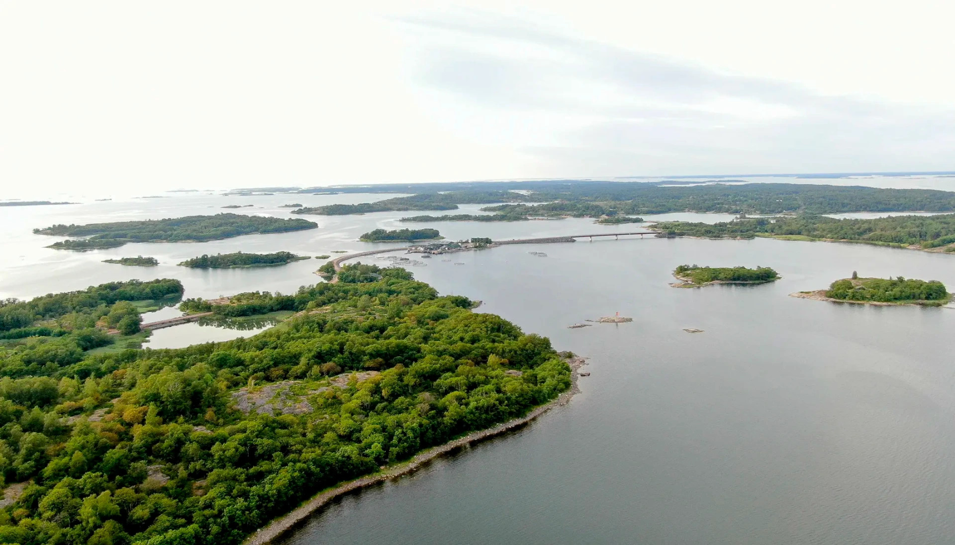 Brändön saaristo on rauhallinen reissukohde.
Ilmakuva laajasta jokimaisemasta, vihreistä saarista ja sillasta, pilvinen taivas ja satama-alue näkyy.