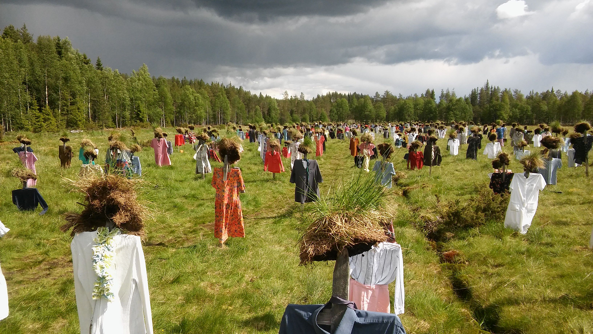Hiljainen kansa, löytyy noin 30 kilometriä Suomussalmelta pohjoisen suuntaan.
Ulkoilmassa metsäniityllä ihmiset seisovat rivissä, yllään värikkäät mekot ja hameet, taustalla pilvinen taivas.