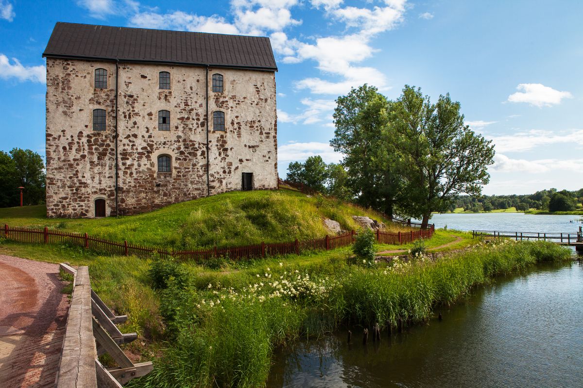 Kastelholman linna on Sundin kunnan tunnetuin vierailukohde.
Kivinen vanha rakennus järven rannalla, vihreä nurmikko ja puinen aita sinisellä taivaalla.