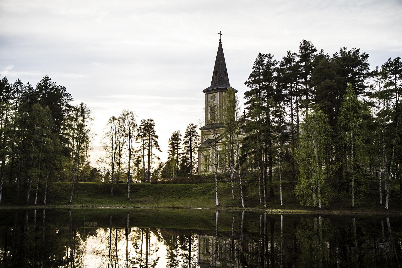 Sotkamon kirkko sijaitsee Sotkamojärven rannalla,
Kirkontorni metsän keskellä, heijastuu rauhalliseen lampeen, taustalla koivuja ja mäntyjä pilvisellä säällä.