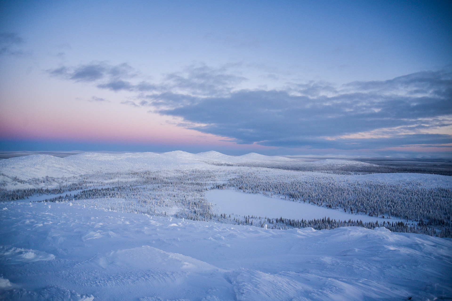 Pyhä-Nattanen. Sodankylä.
Talvimaisema lumisilla rinteillä ja metsillä, sininen taivas ja pilviä, kaukaiset harjanteet hämärässä.