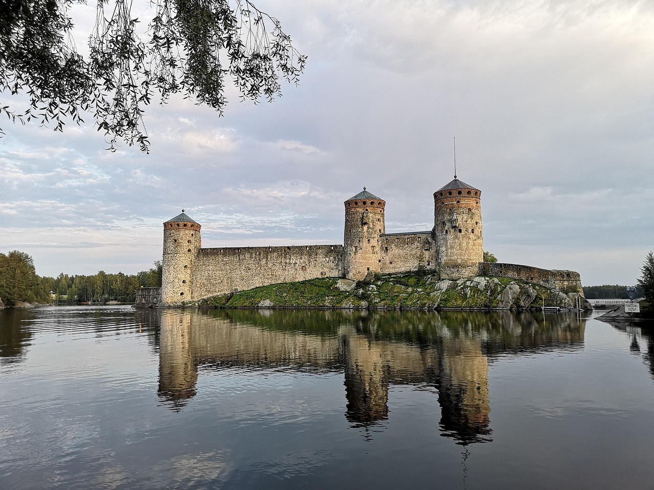 Olavinlinna on Savonlinnan suosituin vierailukohde.
Kivinen linnoitus kolmella tornilla heijastuu järvessä, taustalla pilvinen taivas ja riippuva puunoksisto.