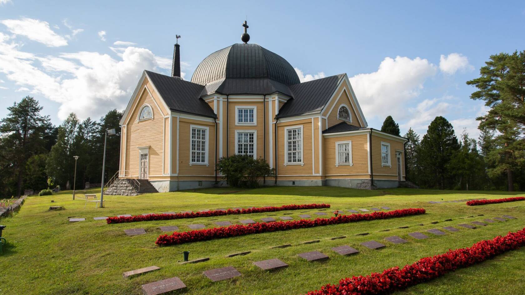 Rääkkylän kirkko on valmistunut vuonna 1851.
Keltainen kirkko, musta katto ja kupoli, punaisia kukkapenkkejä sekä hautakiviä nurmikolla.