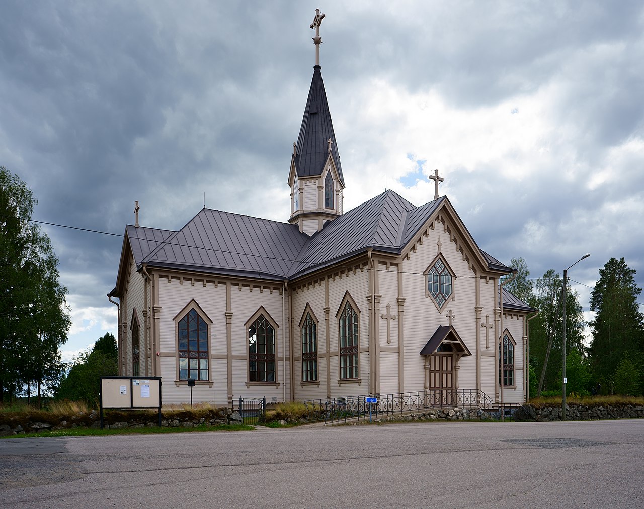 Reisjärven kirkko on rakennettu vuonna 1820.
Valkoinen puukirkko, tummanharjakattoinen torni ja ristejä, pilvinen taivas, edessä asfalttitie ja nurmikko.