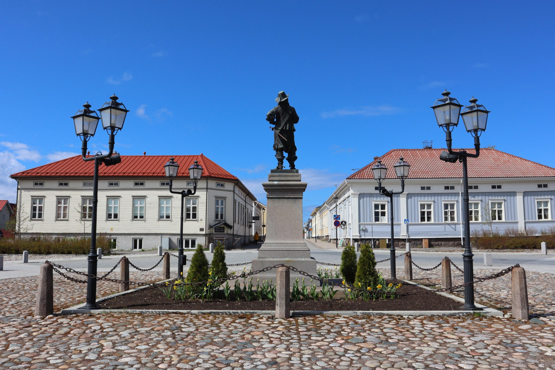 Vanha Raahe on idyllinen puutaloalue.
Kivipatsas jalustalla, ketjuaita ja lyhtypylväät, taustalla valkoiset rakennukset punaisilla katoilla sinisellä taivaalla.