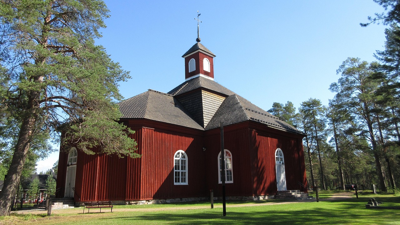 Pudasjärven kaunis puinen kirkko on rakennettu 1781.
Punainen puukirkko, harjakatto ja kellotorni, ympärillä vihreä nurmikko ja korkeat puut sinisellä taivaalla.