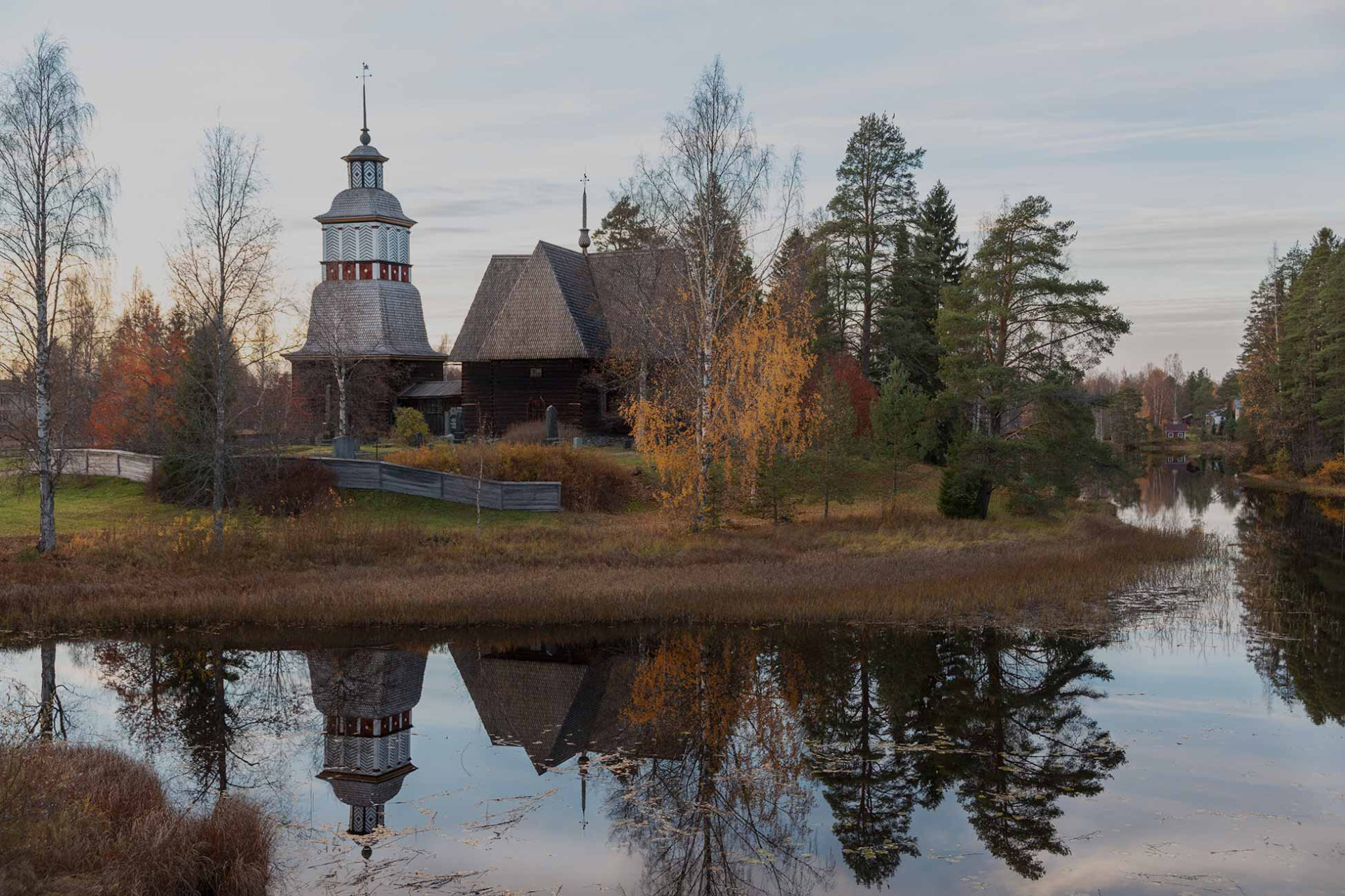 Petäjäveden vanha kirkko on UNESCON maailmanperintökohde.
Puukirkko ja kirkontorni järven rannalla, syksyisiä puita, pilvinen taivas ja heijastus vedessä.