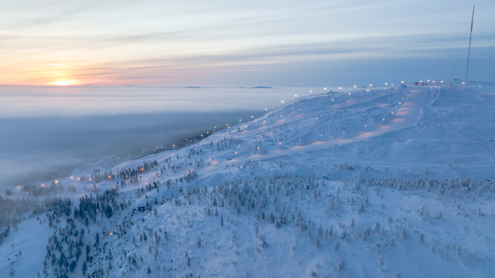 Pyhätunturi on upea talvilomakohde.
Talvinen tunturimaisema, valaistu rinne ja hiihtokeskuksen valaistut polut hämärässä, taustalla meri ja taivas.