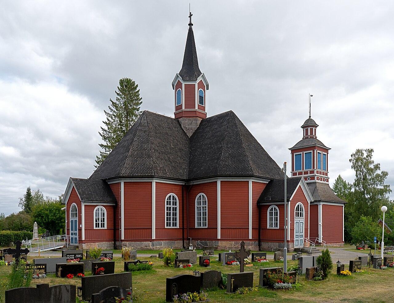Oulaisten kirkko on valmistunut vuonna 1753.
Punainen puukirkko, korkea torni ja hautausmaa, taustalla pilvinen taivas ja vihreitä puita.
