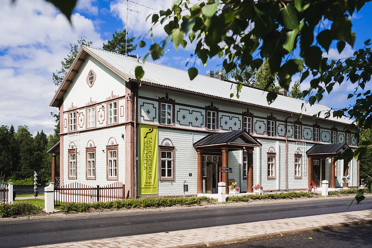 Taidekeskus Salmela Mäntyharjulla.
Valkoinen historiallinen rakennus, jossa on koristeelliset ikkunat, kyltti ja katos, puiden lehdet kehyksessä.