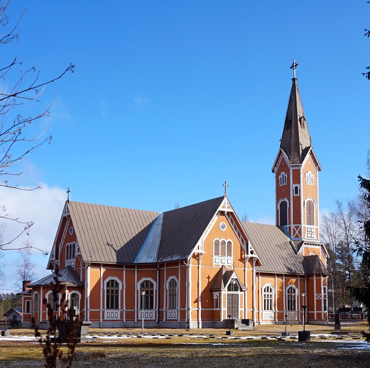 Loviisa Charlottan kirkko Multialla.
Punatiilinen kirkko, korkea kellotorni ja harjakatto, sininen taivas, puiden oksia sivuilla.