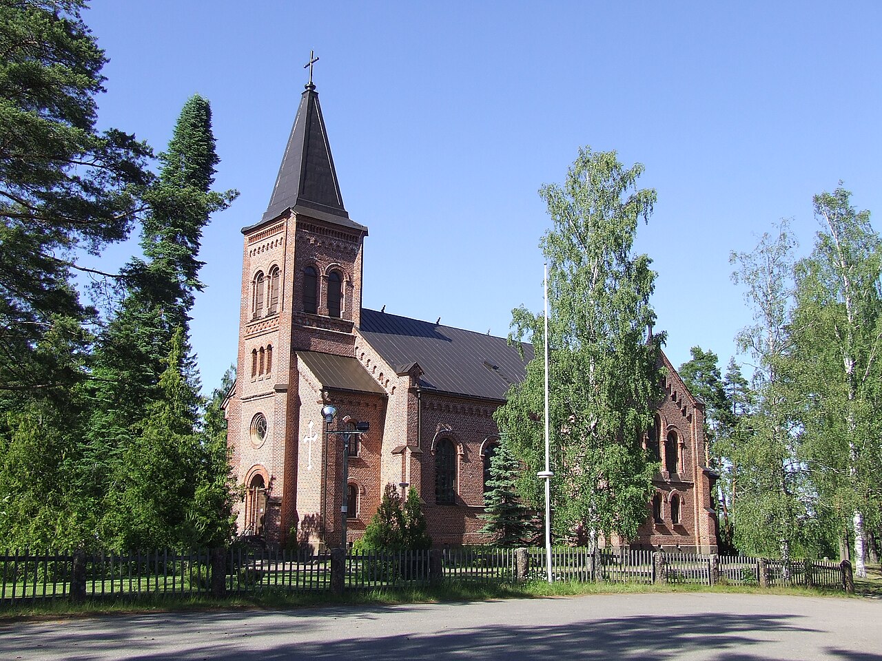 Miehikkälän kirkko on rakennettu vuosina 1879–1881
Punatiilinen kirkko kellotorneineen, ympärillä korkeita puita ja sininen taivas, edessä aita ja tie.