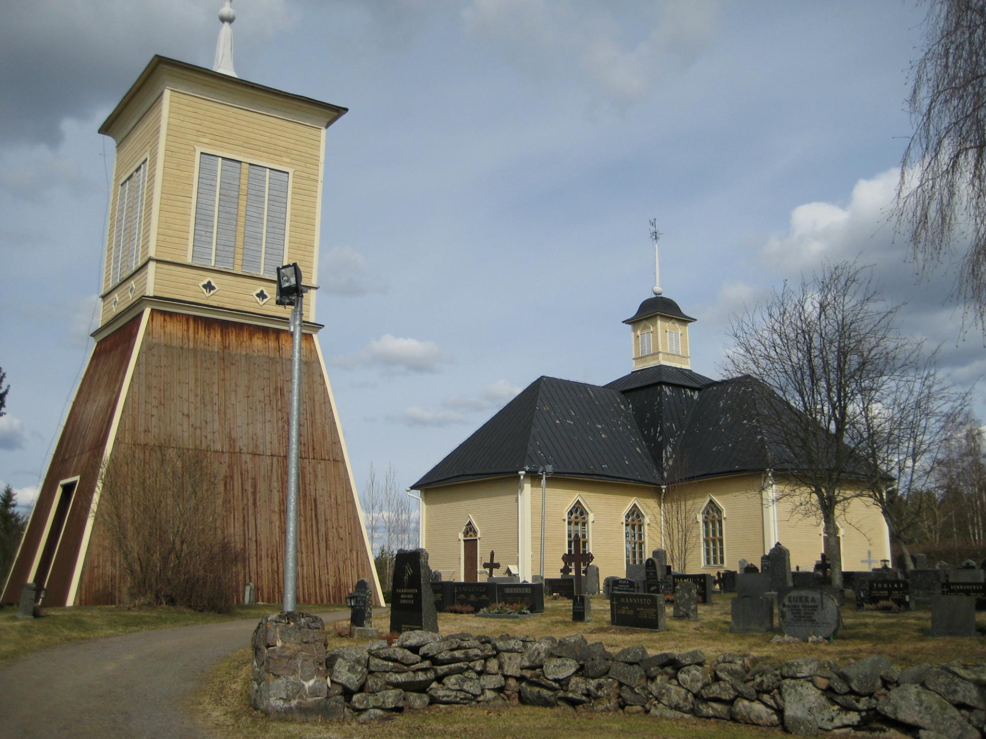 Merijärven Sophia Magdalena -puukirkko ja Merijärven historiallinen kellotapuli.
Kirkko ja puinen kellotorni hautausmaalla, kiviröykkiöitä ja hautakiviä taustalla pilvisellä taivaalla.