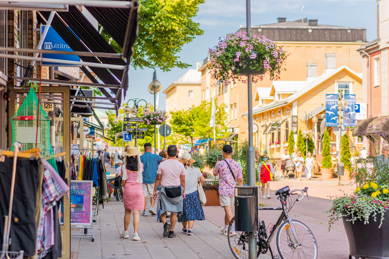 Torggatanin kävelykatu Maarianhaminassa.
Kaupunkikävelykatu, ihmiset kävelevät kukkakoristeisilla terasseilla, taustalla aurinkoisia rakennuksia ja polkupyörä.