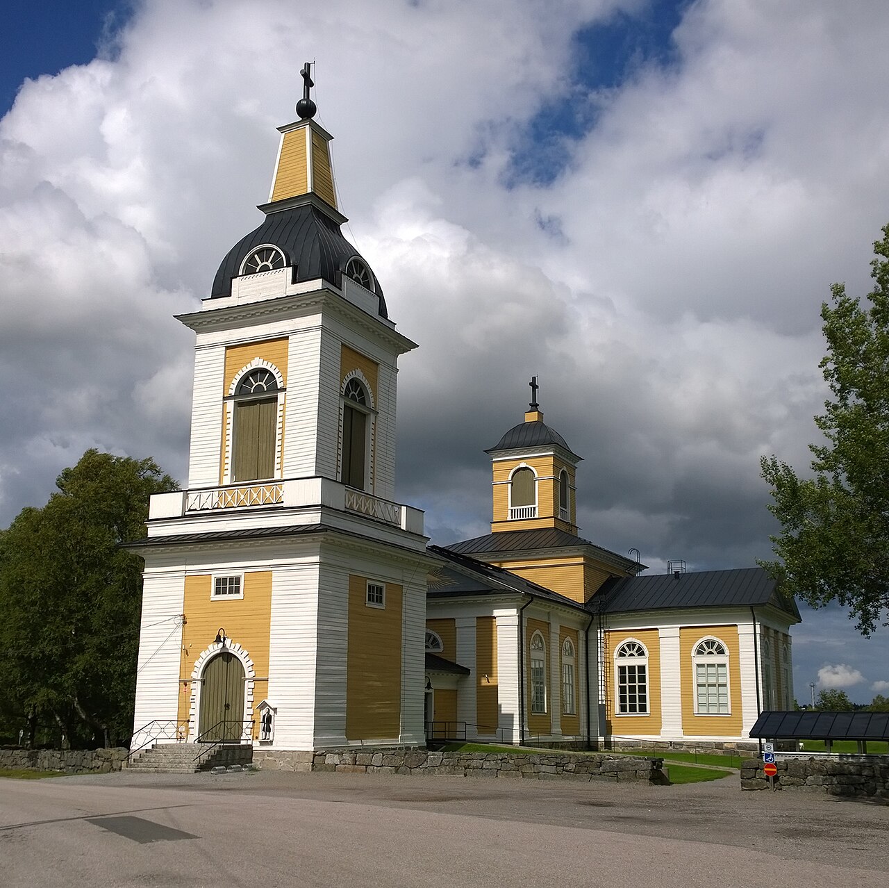 Maalahden kirkko Pohjanmaalla.
Valkoinen ja keltainen kirkko, kellotorni ja kupoli, pilvinen taivas sekä puut ympärillä.
