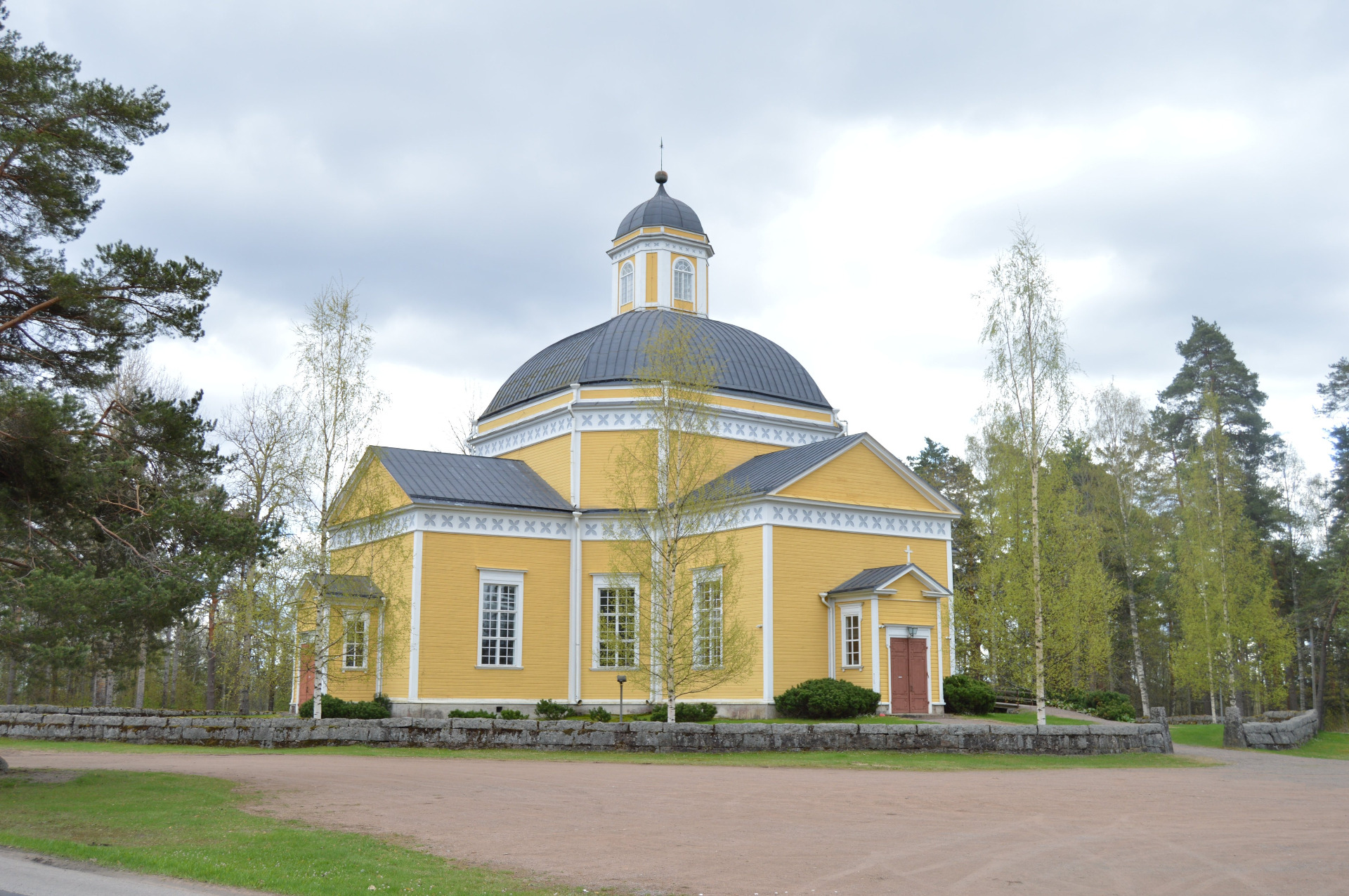 Luumäen kirkko ja historiallinen kirkonmäki on kaunis vierailukohde.
Keltainen kirkko, jossa on kupoli ja ristikko, ympärillä metsää ja koivuja pilvisellä säällä.