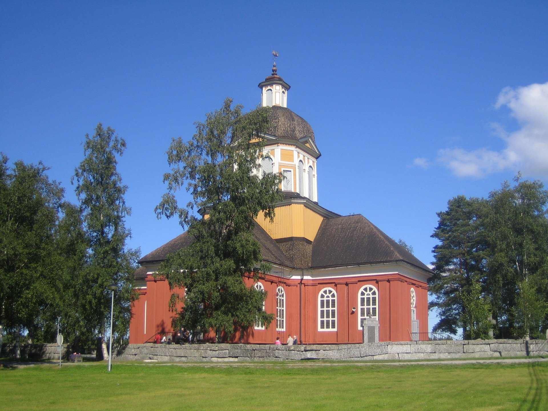 Luodon kirkko Pohjanmaalla.
Punatiilinen kirkko, jossa on sipulikupoli ja kellotorni, ympärillä vihreitä puita sinisellä taivaalla.