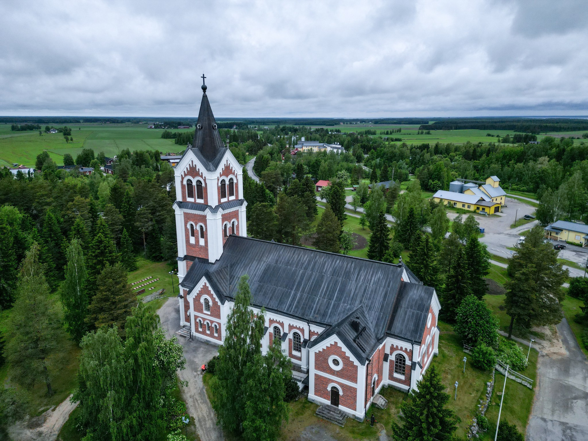 Lumijoen kirkko.
Ilmakuva kirkosta, tiilijulkisivusta ja tornista, ympärillä metsää, peltoja ja pilvinen taivas.