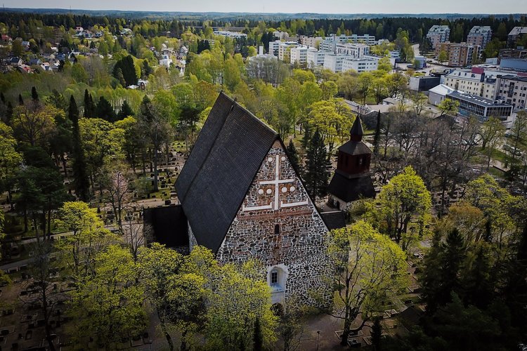 Pyhän Laurin keskiaikainen kirkko Lohjalla.
Ilmakuva tiilirakennuksesta ja kirkon tornista puiden ympäröimänä, taustalla kerrostaloja ja vihreää maisemaa.