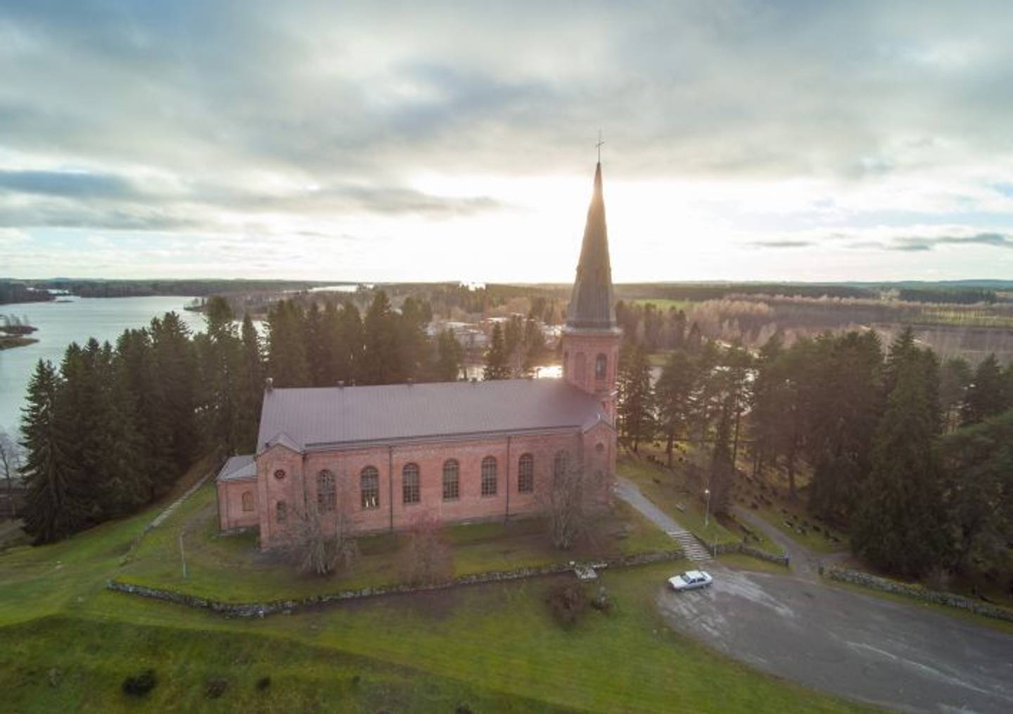 Liperin kirkko Pohjois-Karjalassa
Punatiilinen kirkko kohoaa järven rannalla, korkea torni näkyy auringonpaisteessa puiden ympäröimänä.