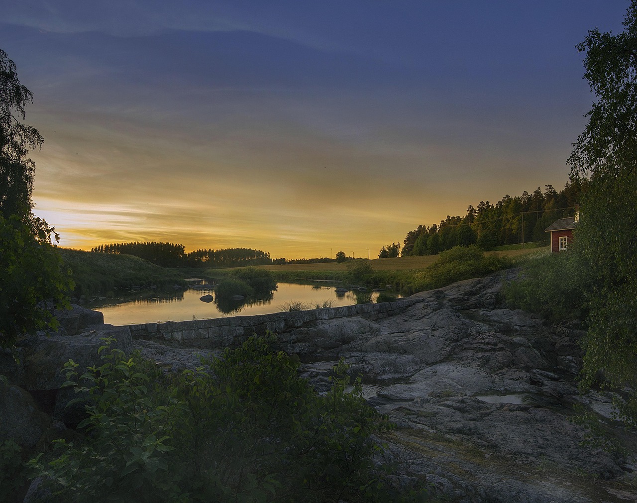 Nautelankoski, Liedon kunnassa.
Auringonlaskun aikaan rauhallinen joki, kivinen ranta, metsää ja pieni punainen mökki taustalla.
