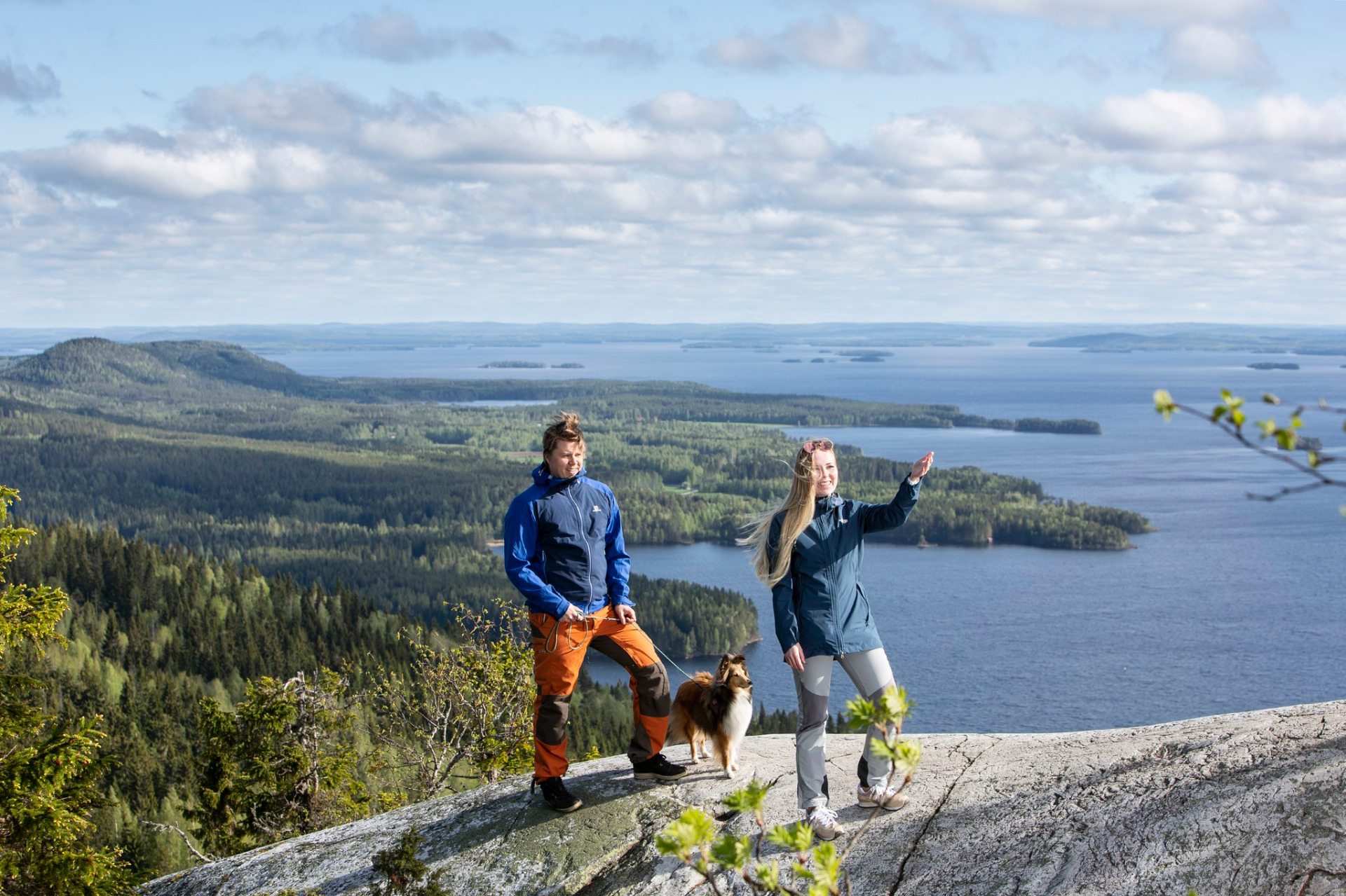 Ukko-Kolin kansallismaisema Pieliselle.
Retkeilijät ja koira kallion reunalla, taustalla järvimaisema, metsät ja saaret pilvisellä taivaalla.