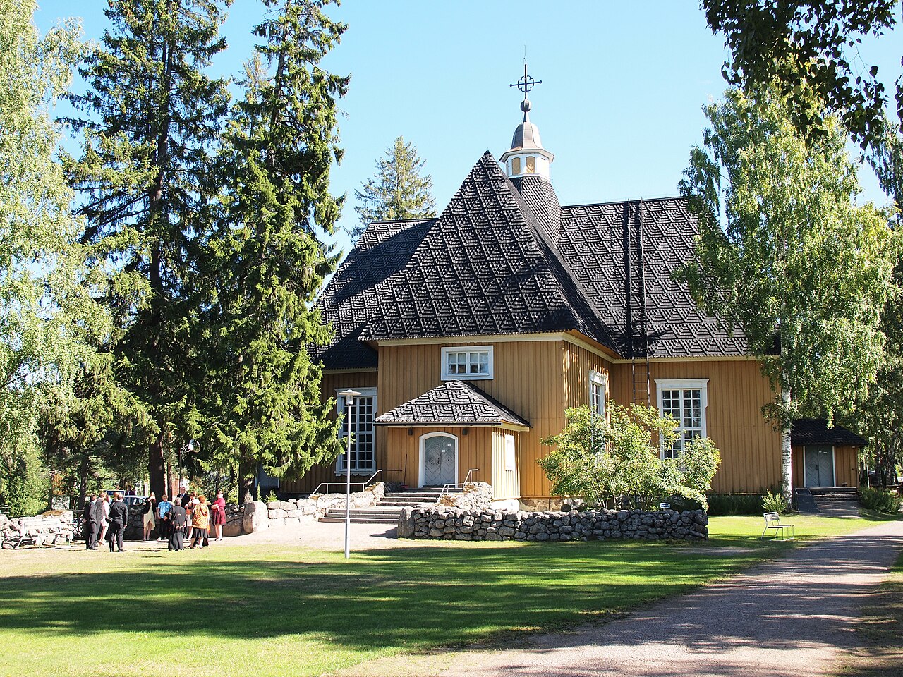Lemin kirkko Etelä-Karjalassa.
Ruskea puukirkko, korkea harjakatto ja ristikruunu, ympärillä vihreitä puita ja nurmikkoa.