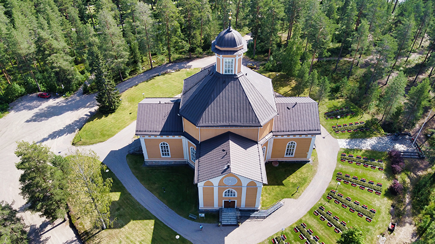 Laukaan kirkko Keski-Suomessa.
Ilmakuva historiallisesta kirkosta, ympärillä vehreä metsä, piha ja rivissä olevia pysäköintipaikkoja.