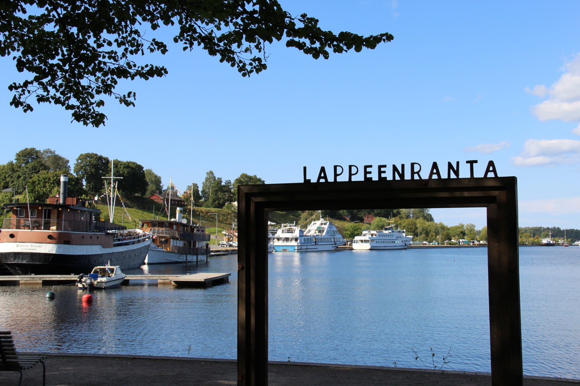 Lappeenrannan rantapuisto.
Satamajoki ja laiturit, taustalla veneitä, ja musta portti, jossa teksti LAPPENRANTA.