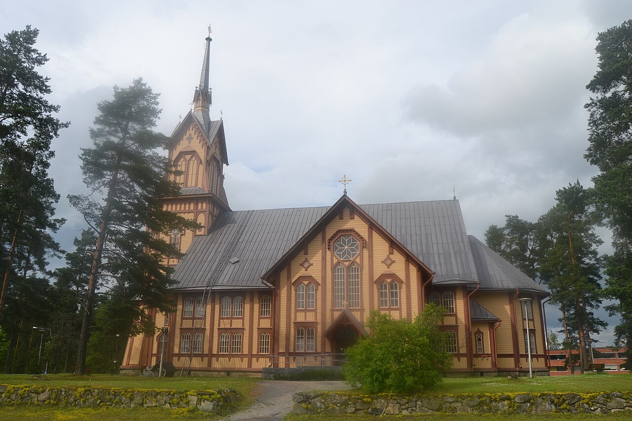 Lapinlahden kirkko Pohjois-Savossa.
Puinen kirkko, korkea kellotorni ja harmaa katto, ympärillä metsää ja pilvinen taivas.