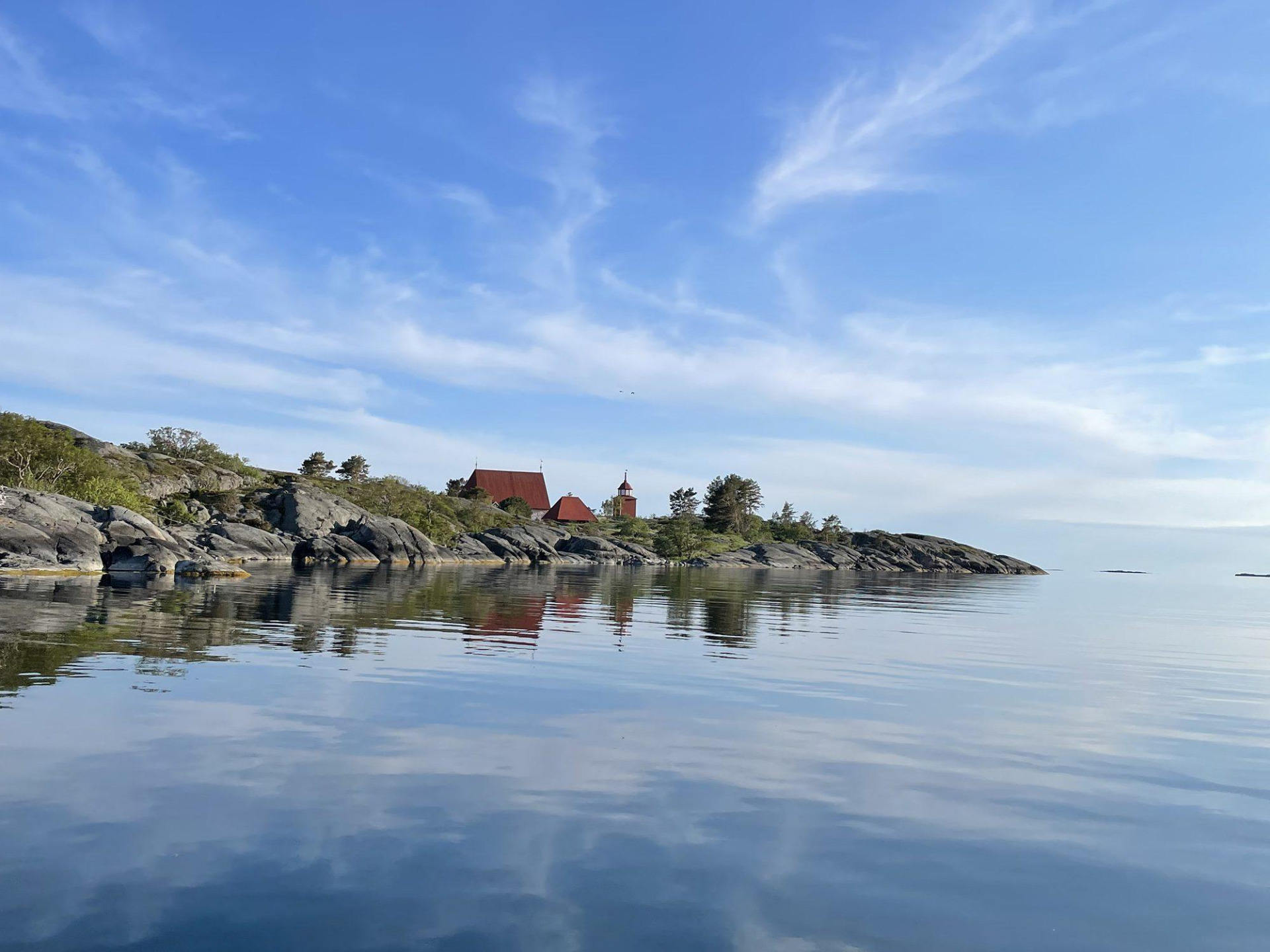 Källskärin rantakalliot Kökarilla.
Punakattoinen kirkko ja kellotorni heijastuvat tyynessä rannikkovedessä, kallioinen saari ja sininen taivas.