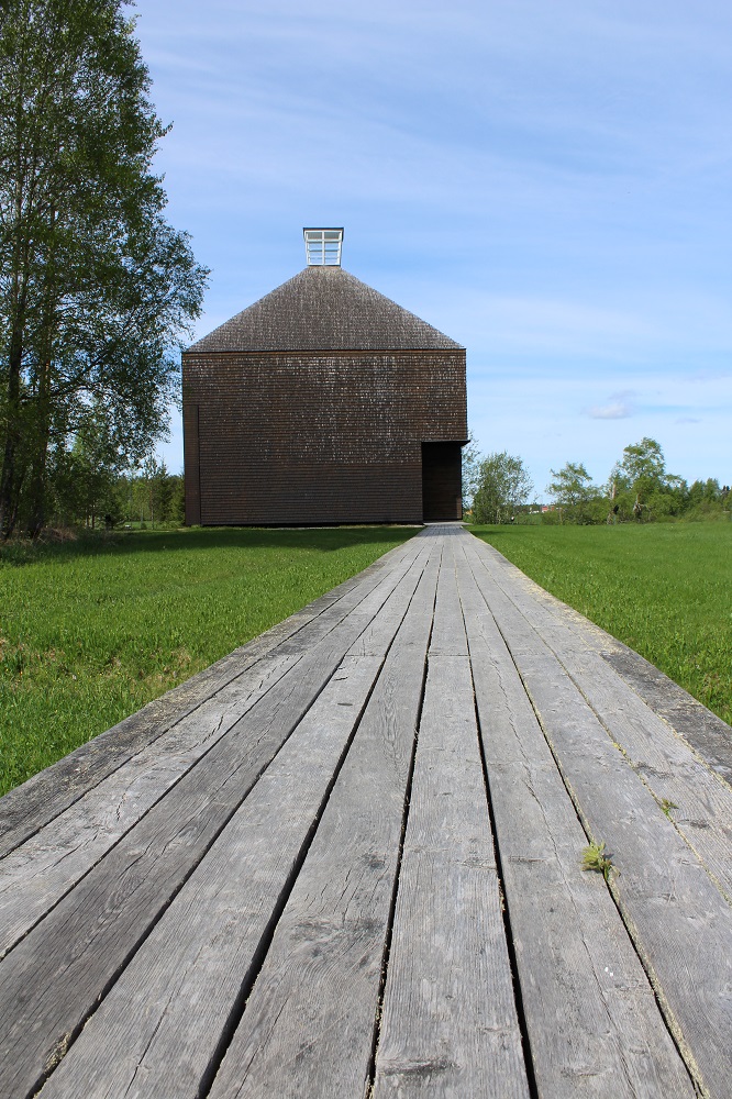 Kärsämäen Paanukirkko Pohjois-Pohjanmaalla.
Harmaa puinen polku johtaa tummanruskeaan lato-rakennukseen vihreällä niityllä, taustalla puita ja sininen taivas.