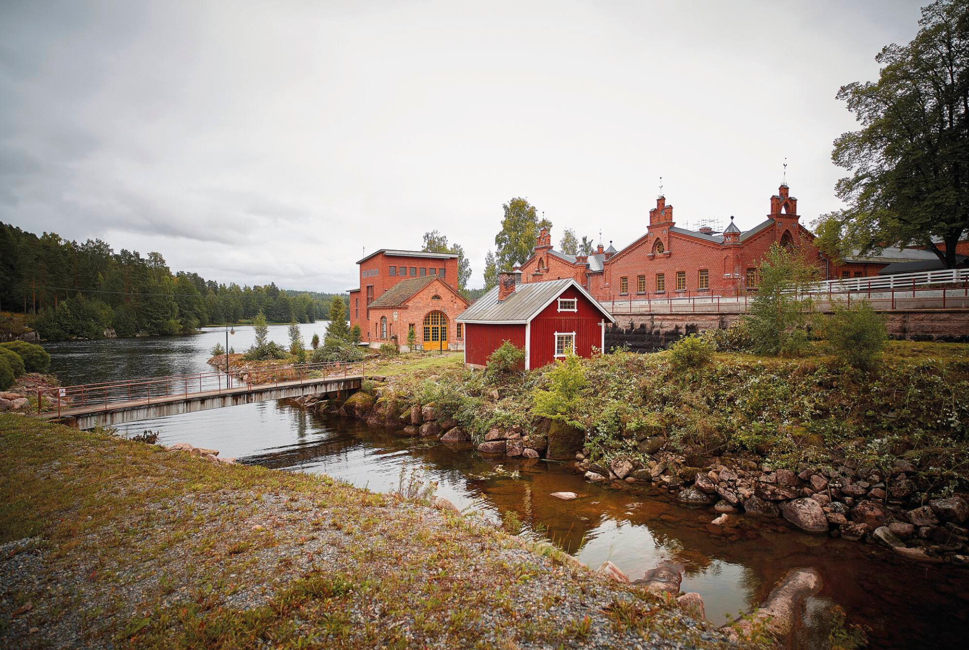Verlan puuhiomo ja pahvitehdas on Kouvolan merkittävin kulttuurihistoriallinen nähtävyys.
Punatiilinen tehdasrakennus ja punainen varastorakennus järven rannalla, puusilta ja pilvinen taivas.