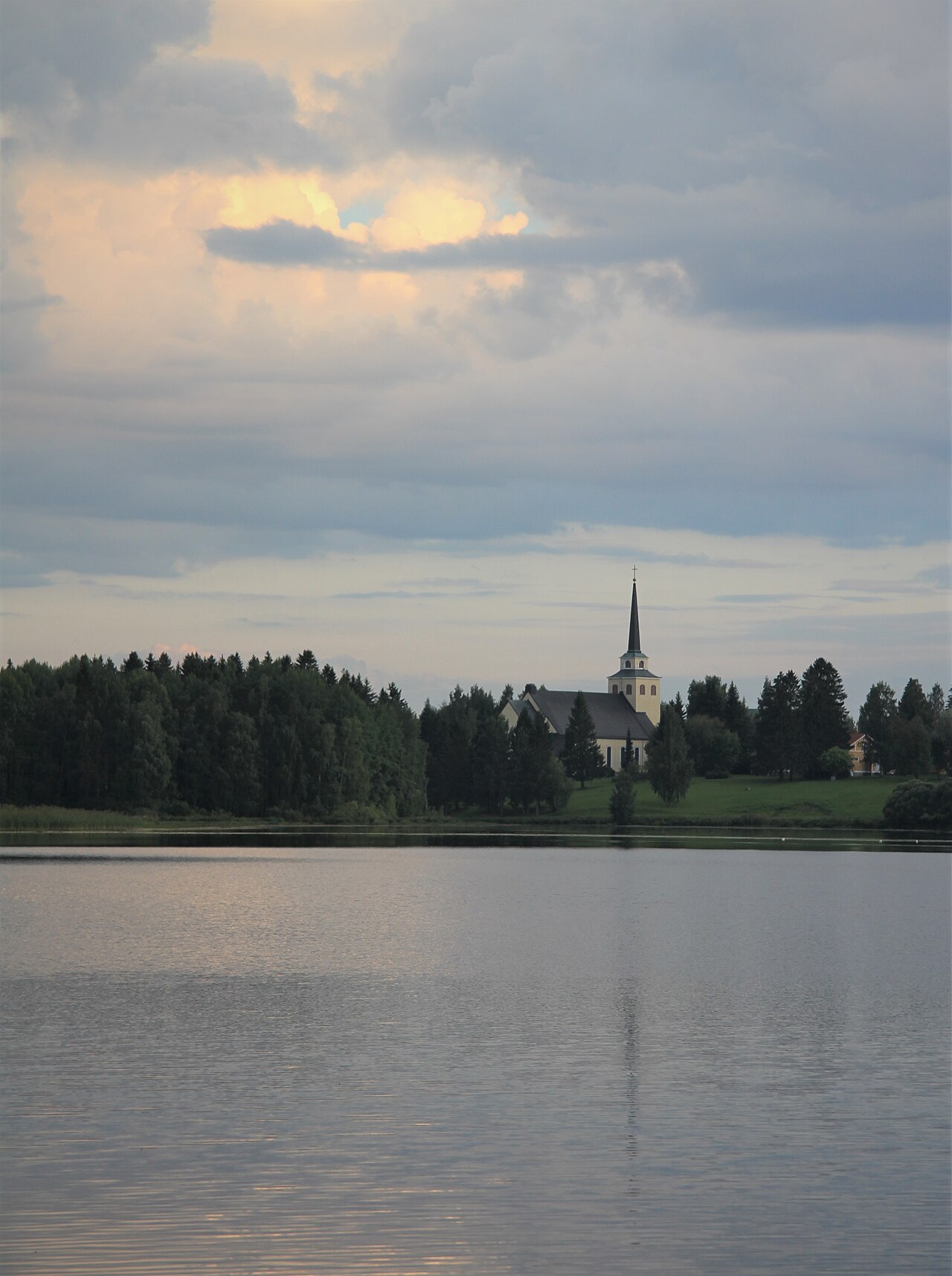 Kiuruveden evankelis-luterilainen kirkko.
Järvimaisema, metsää ja valkoinen kirkko kellotorneineen, taivaalla pehmeitä pilviä auringonlaskun valossa.