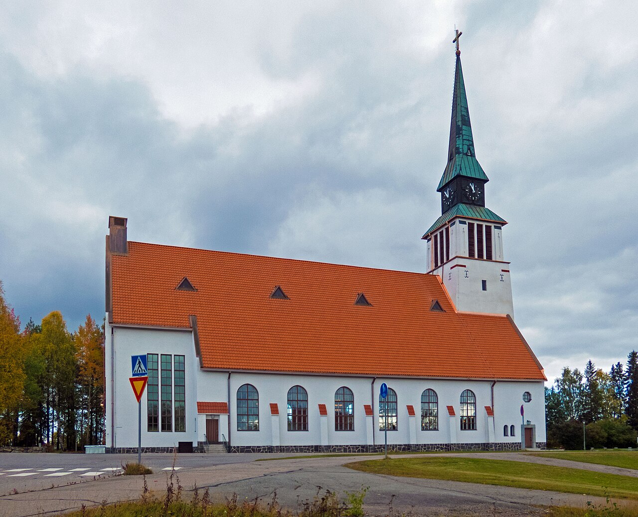 Kemijärven kirkko Lapissa.
Valkoinen kirkko punaisella tiilikatolla ja vihreällä tornikupolilla, pilvinen taivas ja puisto ympärillä.
